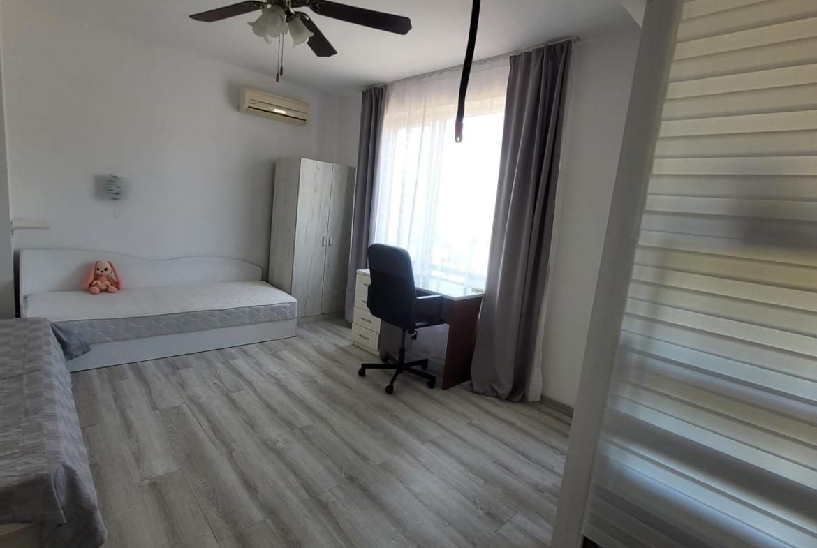 Светлая комната с односпальной кроватью, рабочим столом и шкафом, окна выходят на улицу, стильный интерьер для аренды в Болгарии.