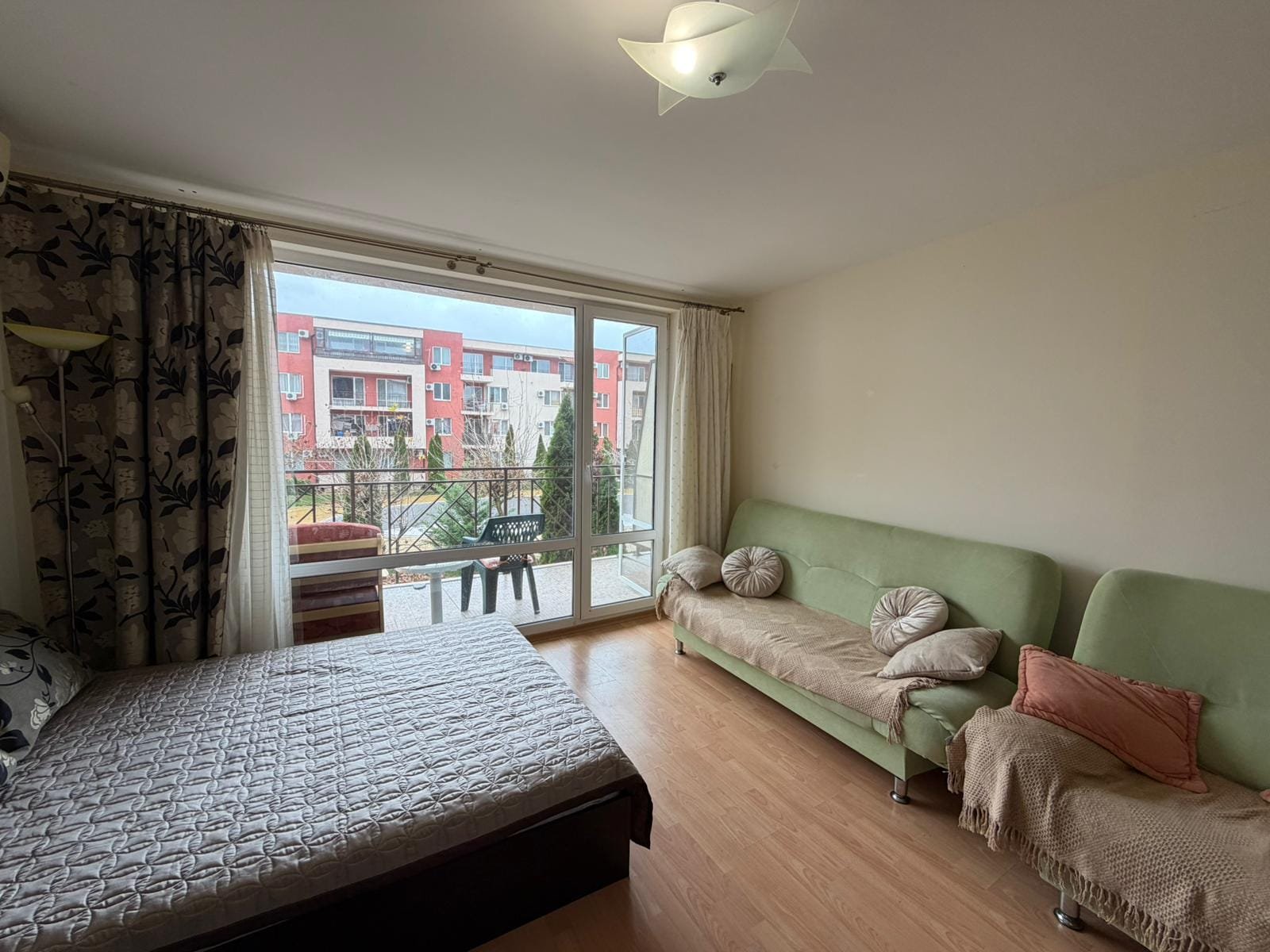 Современный интерьер квартиры с видом на улицу Светлая комната с балконом, кроватью и диваном, идеально подходит для отдыха и жизни в Болгарии.