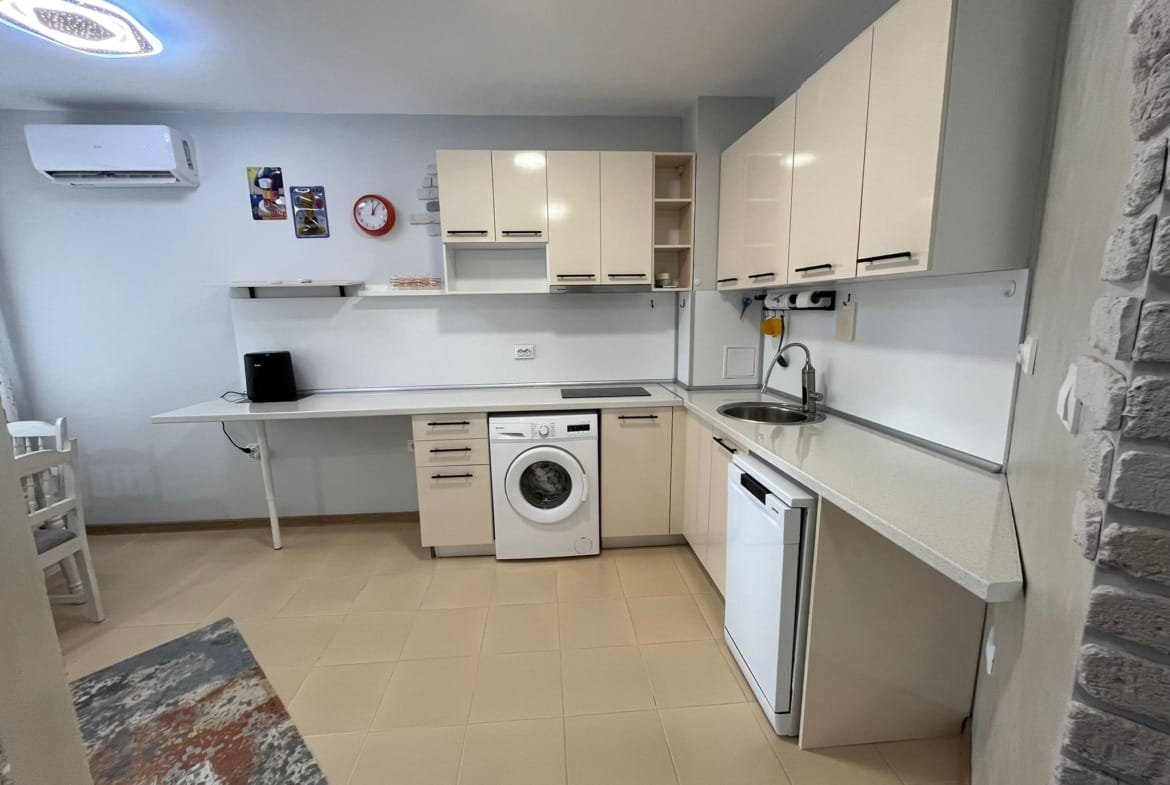 Современная кухня с белыми шкафами, техникой и стиральной машиной, создающая уютную атмосферу для продажи недвижимости в Болгарии.