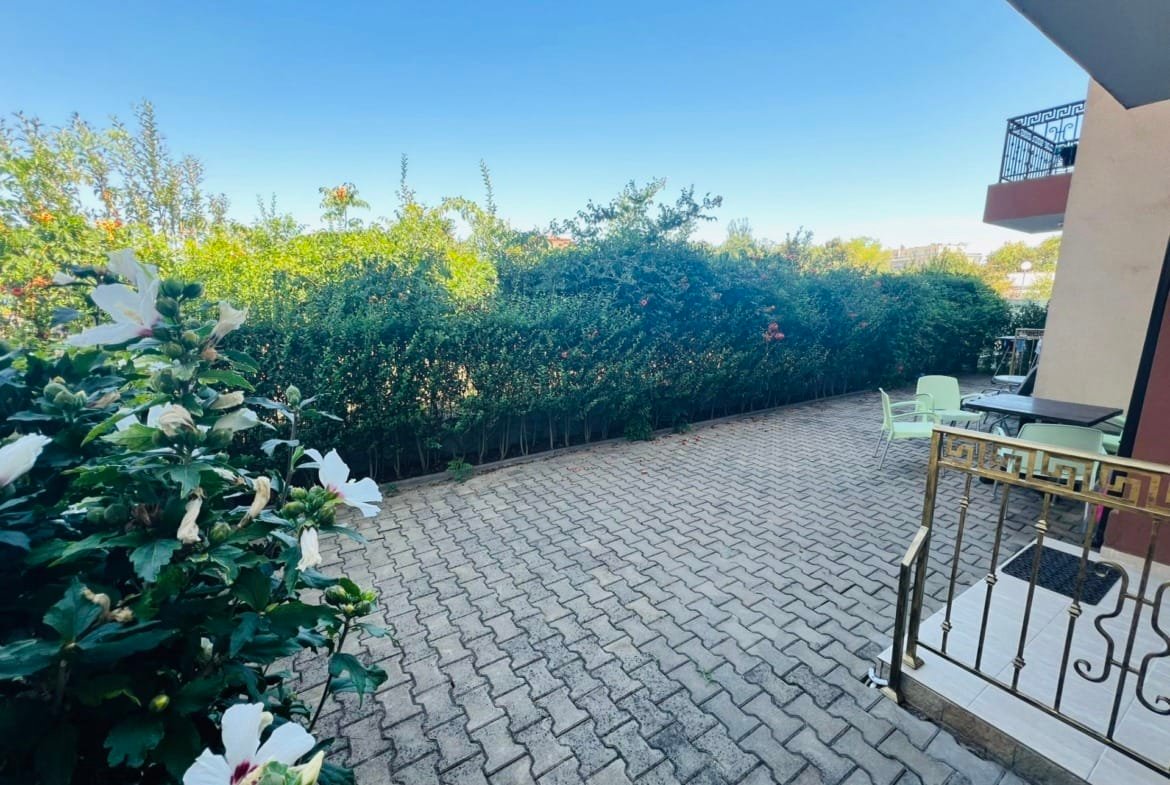 Просторная терраса с кирпичной плиткой, зелеными кустами и столиками для отдыха в районе продажи недвижимости в Болгарии.