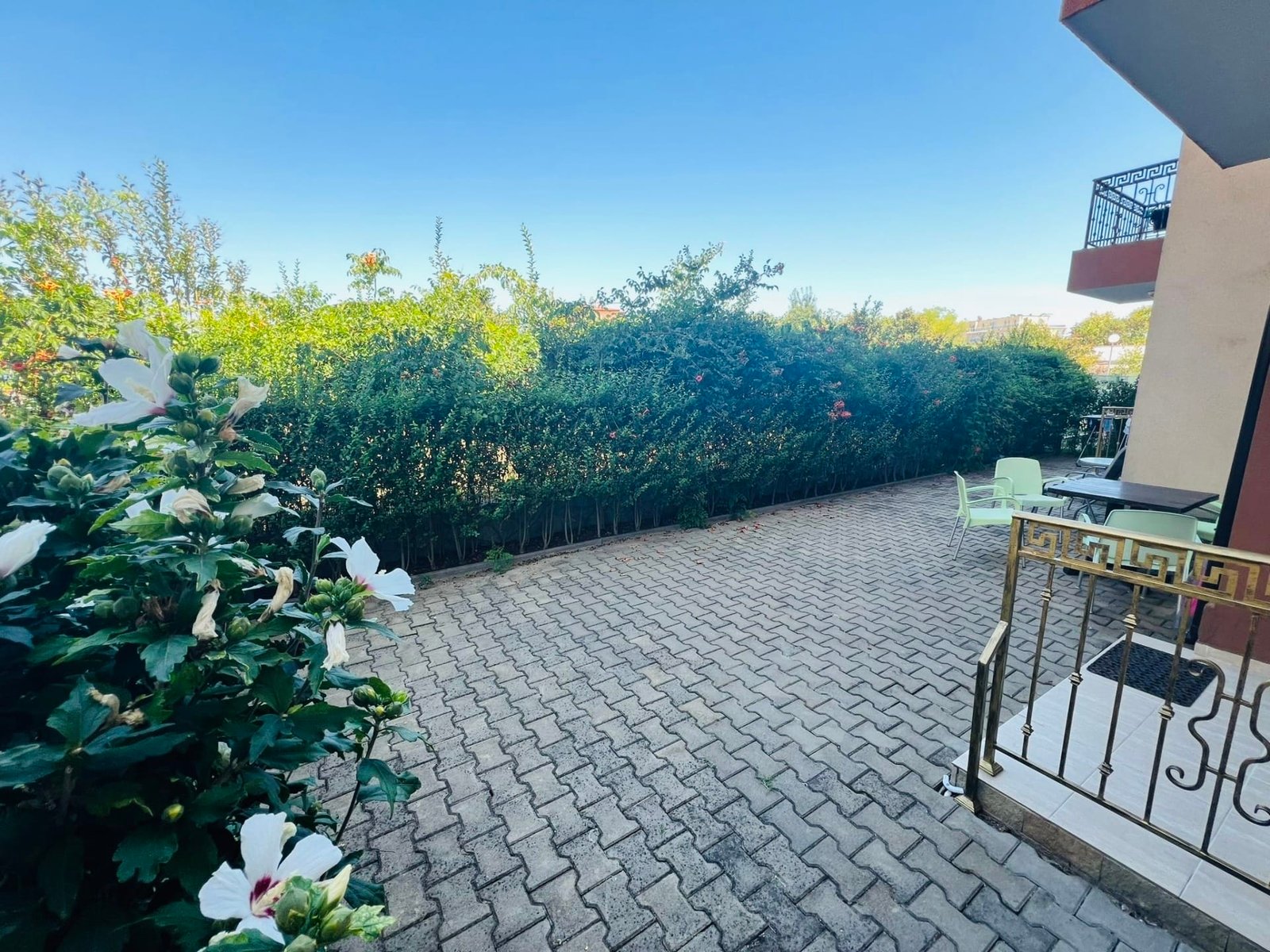 Просторная терраса с кирпичной плиткой, зелеными кустами и столиками для отдыха в районе продажи недвижимости в Болгарии.