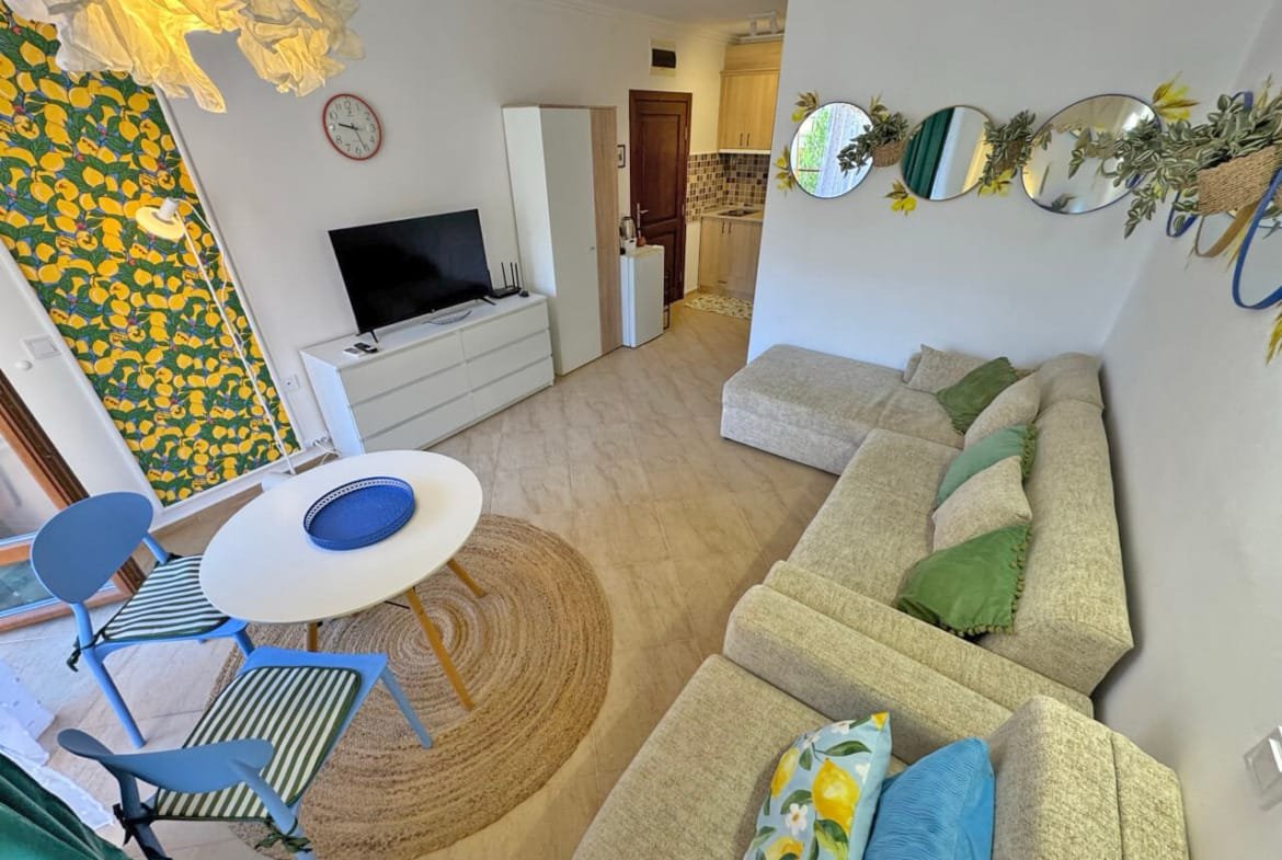 Современная гостиная с диваном, круглым столом и телевизором, оформленная в ярких цветах, идеальная для квартиры у моря в Болгарии.