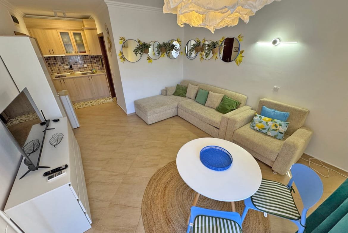 Современная гостиная с угловым диваном, круглым столом и телевизором, кухня в фоне, зелёные и бежевые оттенки, идеально для жилья в Болгарии.