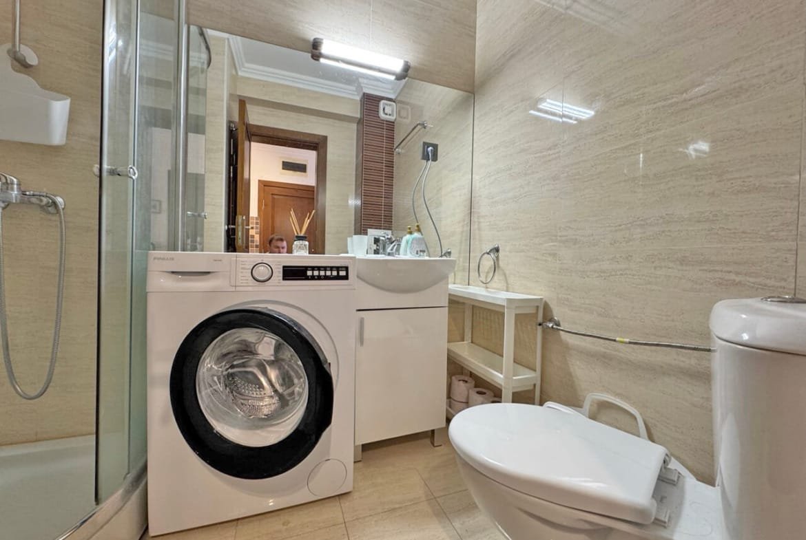 Современная ванная комната с душевой кабиной, стиральной машиной и раковиной, выполненная в светлых тонах, идеально подходит для апартаментов в Болгарии.