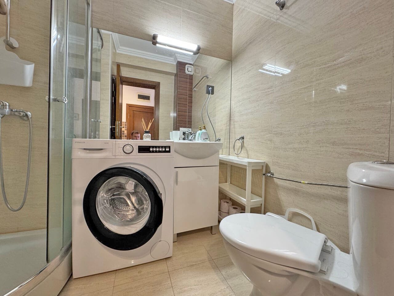 Современная ванная комната с душевой кабиной, стиральной машиной и раковиной, выполненная в светлых тонах, идеально подходит для апартаментов в Болгарии.