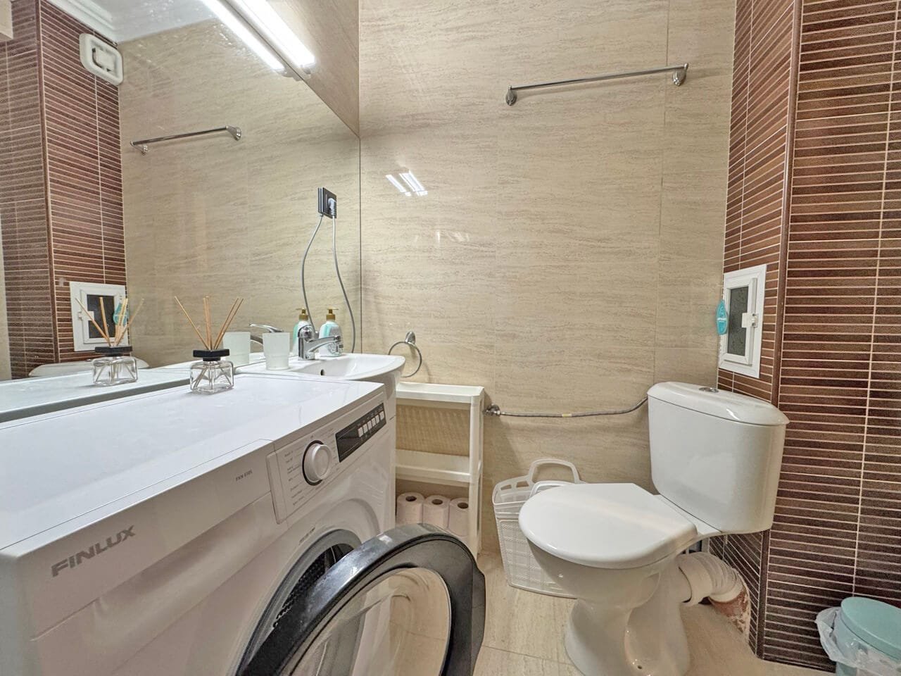 Современная ванная комната с прачечной, стиральной машиной, плиткой бежевых и коричневых оттенков, светлая и функциональная.