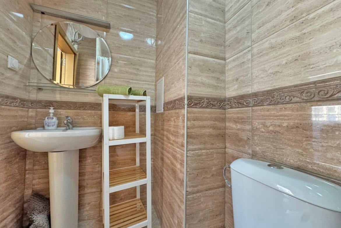 Современная ванная комната с умывальником, зеркалом, туалетом и открытыми полками, отделка в светлых тонах.