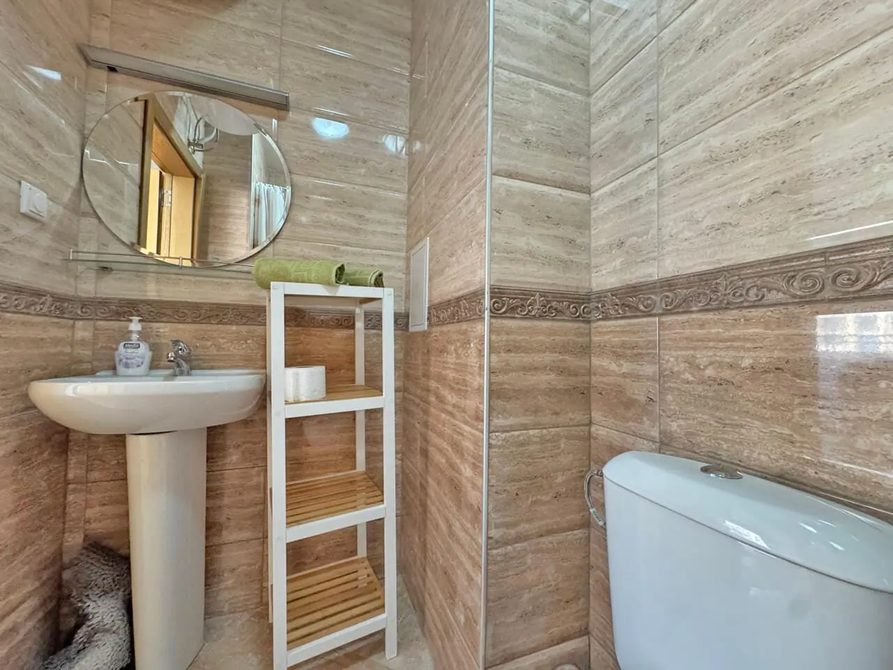 Уютная ванная комната с современным дизайном и бытовыми принадлежностями Современная ванная комната с умывальником, зеркалом, туалетом и открытыми полками, отделка в светлых тонах.