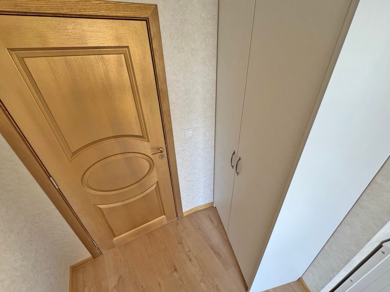 Деревянная дверь и шкаф в интерьере квартиры. Элегантная входная дверь с золотистой отделкой, рядом шкаф для хранения, теплый пол, светлые тона интерьера.