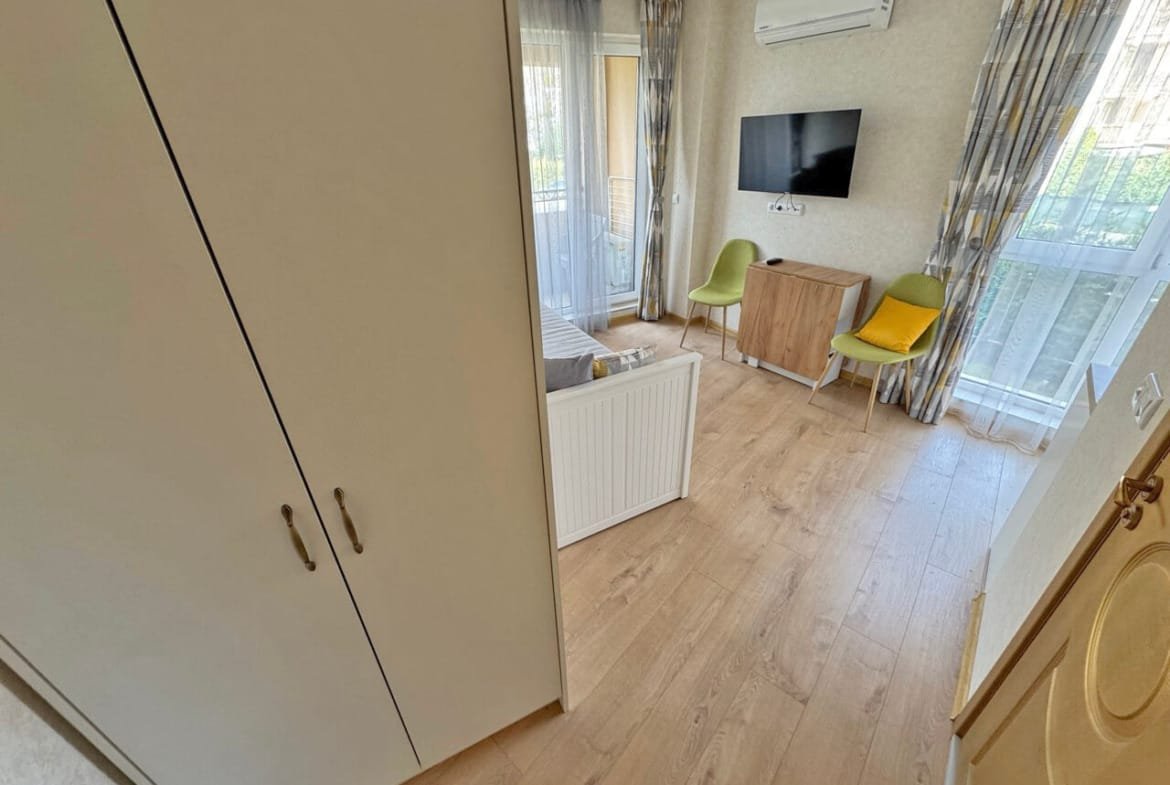 Современная гостиная с телевизором и балконом, светлая отделка, удобная мебель, квартира в Болгарии, идеальна для отдыха у моря.