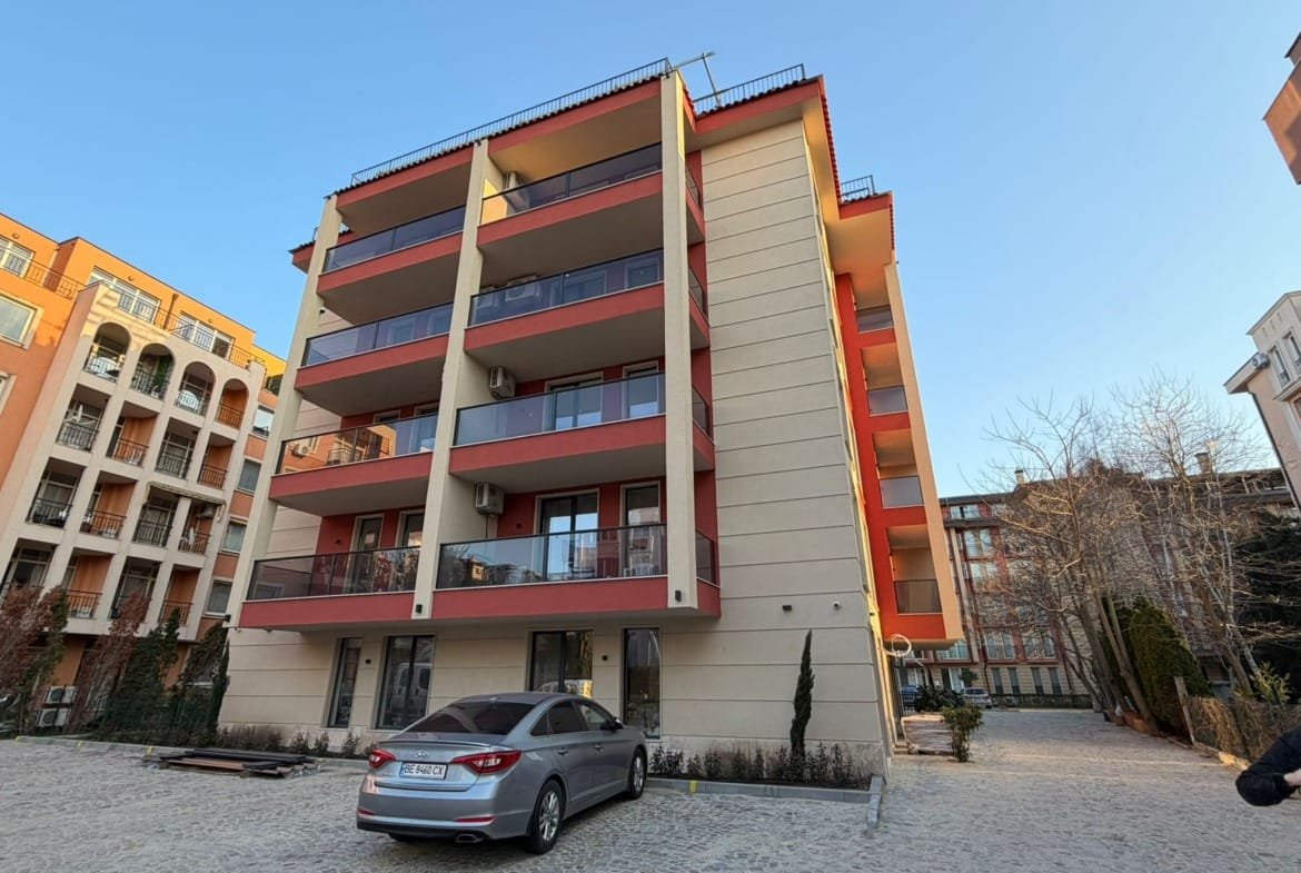 Современное жилое здание с балконами в Болгарии, окруженное парковкой и зелеными насаждениями. Подходит для покупки недвижимости.