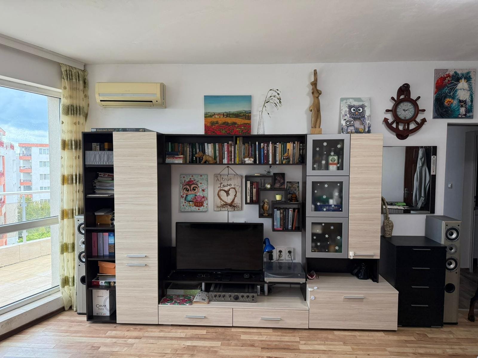 Современный интерьер квартиры в Болгарии с телевизором, книжными полками и яркими художественными деталями.