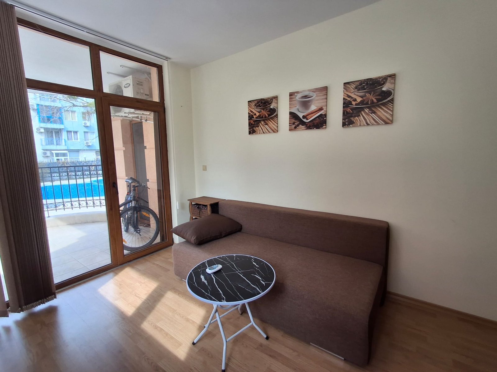 Уютная гостиная с диваном, круглым столиком и окнами, выходящими на балкон с видом на бассейн.