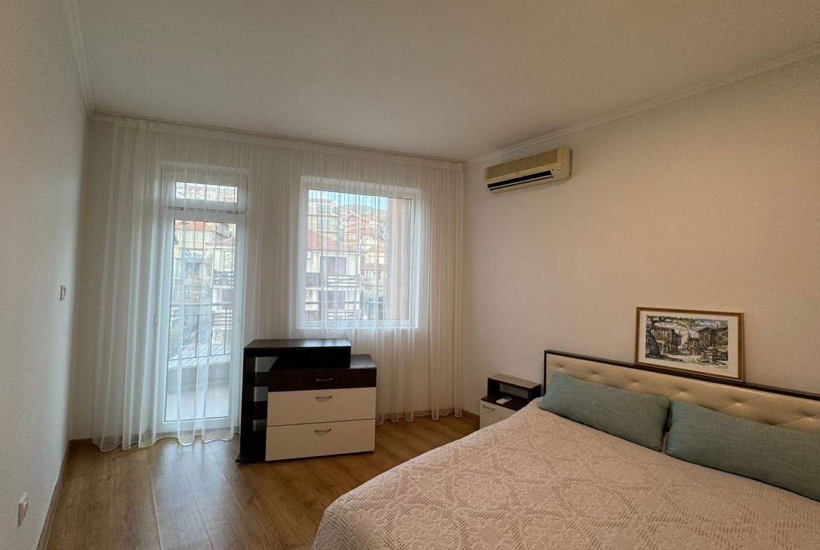 Современная спальня с двуспальной кроватью, комодом и оконными шторами в светлых тонах, подходит для покупки недвижимости в Болгарии.