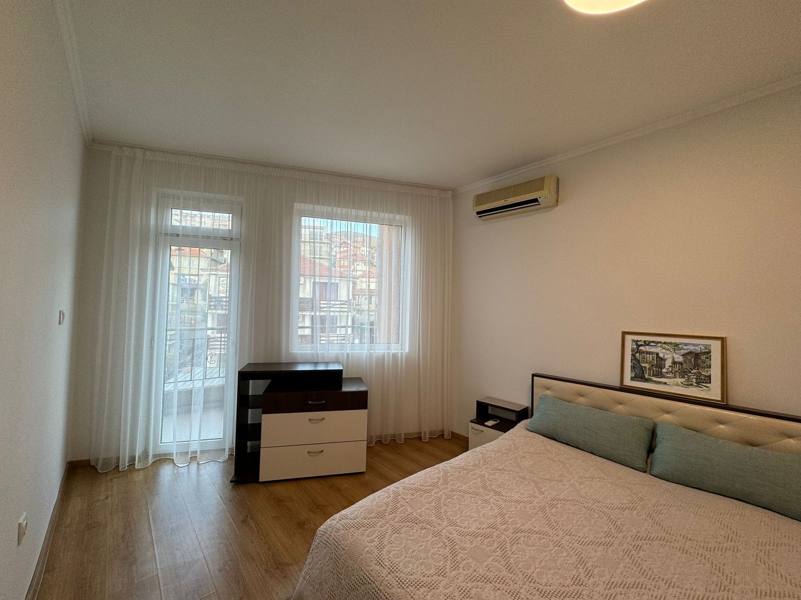 Современная спальня с балконом и светлыми шторами Современная спальня с двуспальной кроватью, комодом и оконными шторами в светлых тонах, подходит для покупки недвижимости в Болгарии.