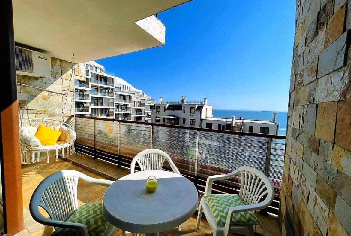 Балкон с белыми креслами и круглым столом, вид на море и жилые комплексы в Болгарии, идеальное место для отдыха.