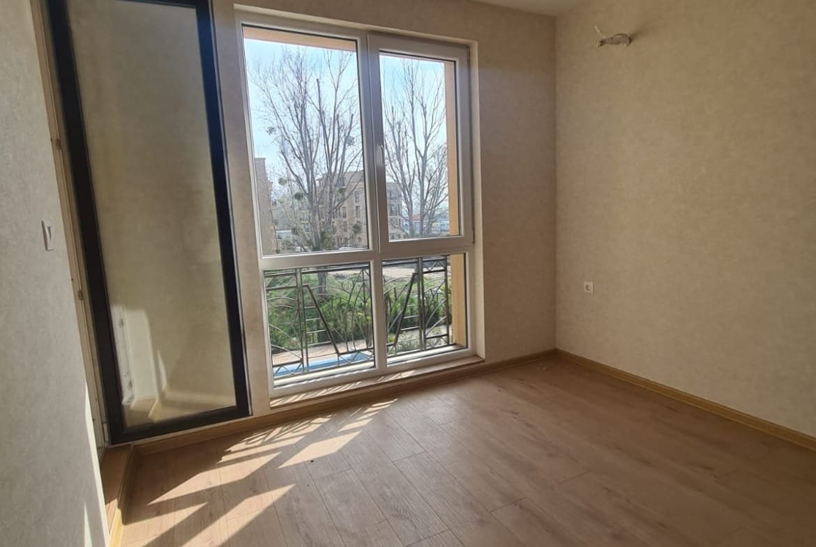 Светлая комната с большими окнами, выходящими на улицу, с деревянным полом и нейтральными стенами, готовая к продаже в Болгарии.