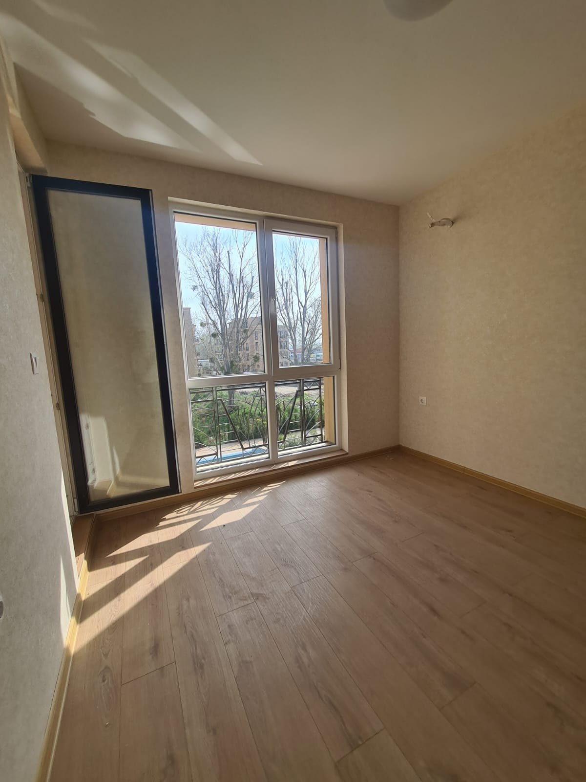 Светлая комната с большими окнами, выходящими на улицу, с деревянным полом и нейтральными стенами, готовая к продаже в Болгарии.