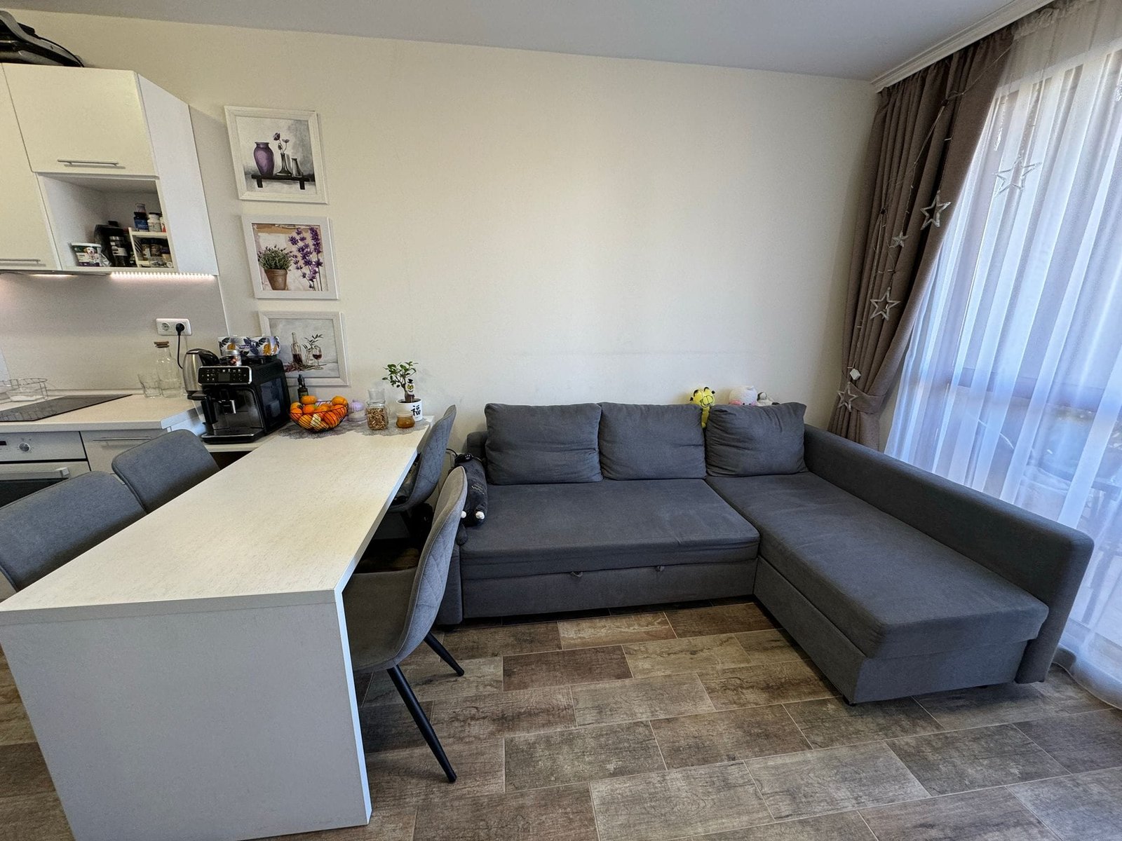 Современная гостиная с угловым диваном и кухонным столом Современная гостиная с серым диваном, обеденным столом, кухонной техникой и яркими декоративными элементами в интерьере.