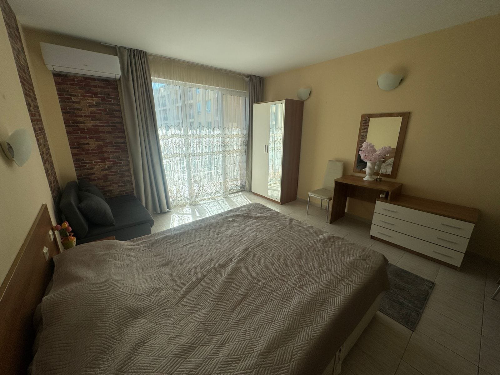 Современная спальня с двуспальной кроватью, комодом и зеркалом, яркое естественное освещение, уютная обстановка для отдыха.