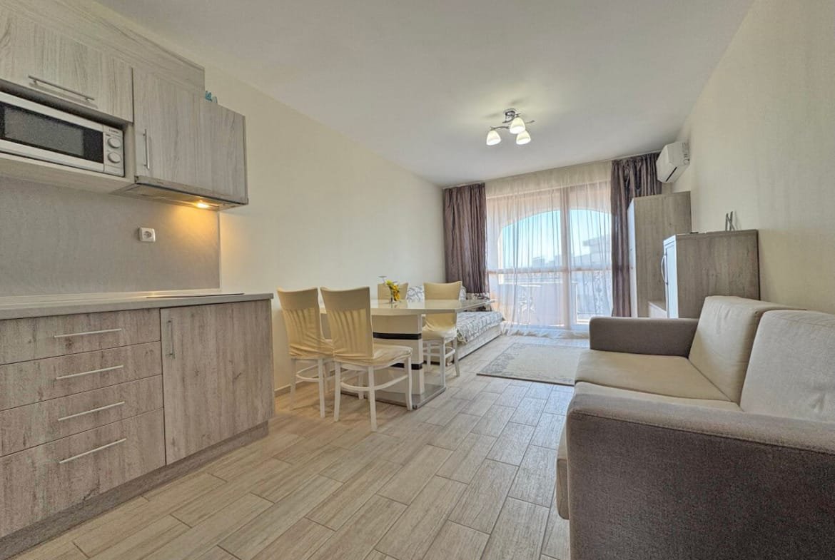 Современная светлая кухня-гостиная с диваном, обеденной зоной и выходом на балкон, оформленная в нейтральных тонах.