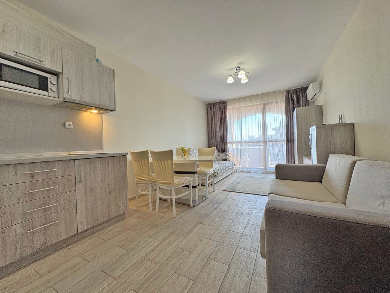 Современная светлая кухня-гостиная с диваном, обеденной зоной и выходом на балкон, оформленная в нейтральных тонах.