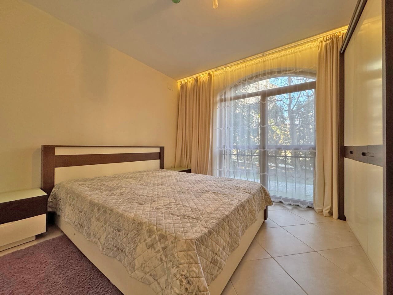 Светлая спальня с двуспальной кроватью, окнами до пола и мягкими шторами, создающими уютную атмосферу.