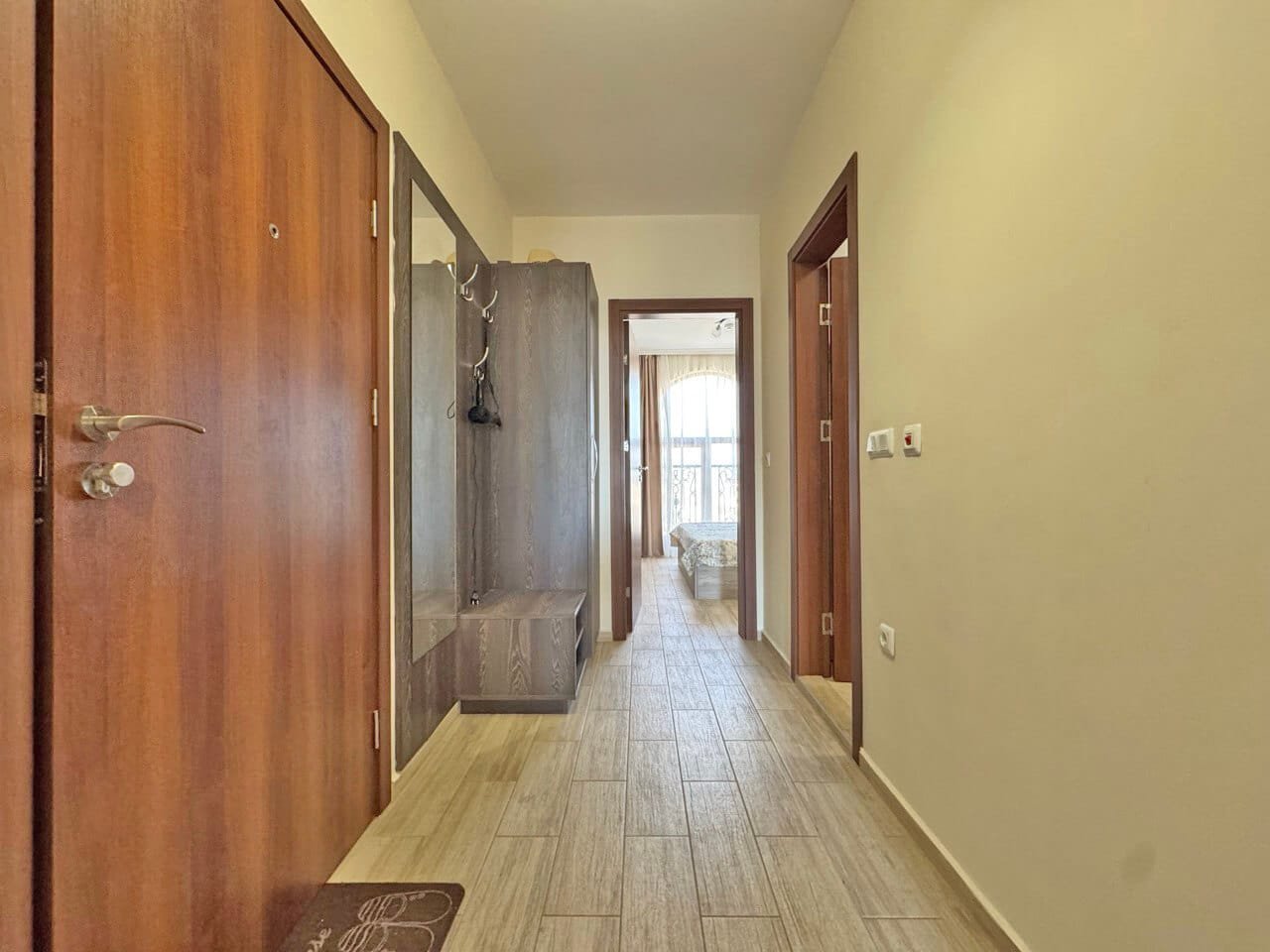 Светлый коридор с деревянной дверью, гардеробом и дверями в комнаты, ведющими к светлой спальне.