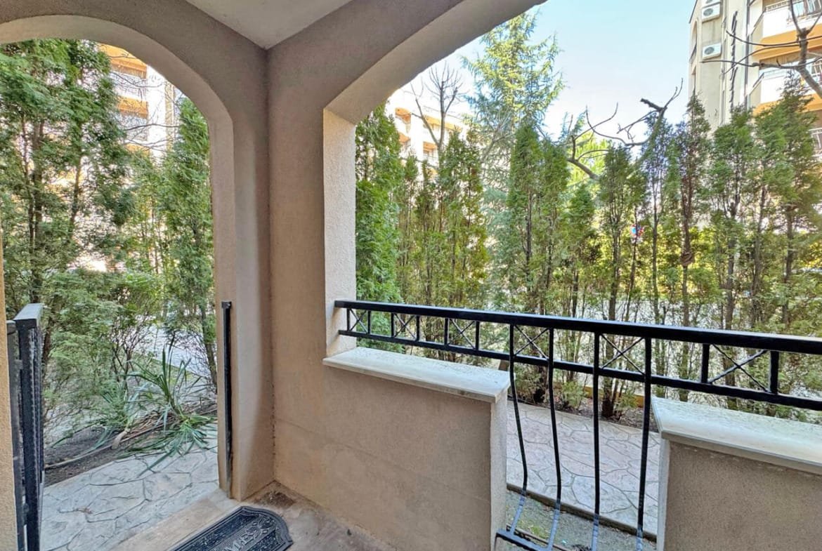 Вид из квартиры на зелёный двор с кустами и тротуаром, арка и балконные перила создают уютную атмосферу.