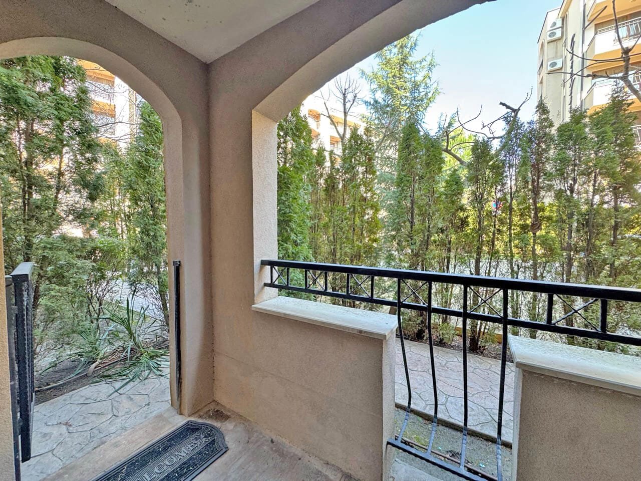 Вид из квартиры на зелёный двор с кустами и тротуаром, арка и балконные перила создают уютную атмосферу.