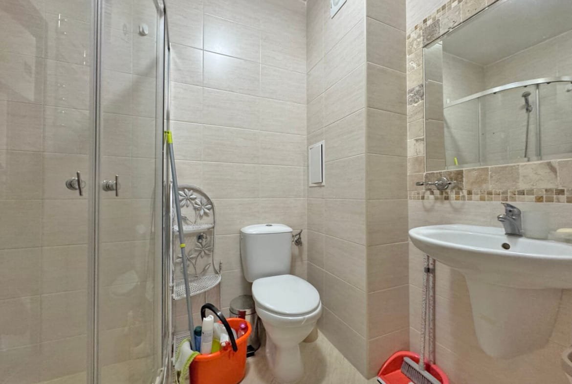 Современная ванная комната с душевой кабиной, унитазом, раковиной и уборочными принадлежностями в углу.