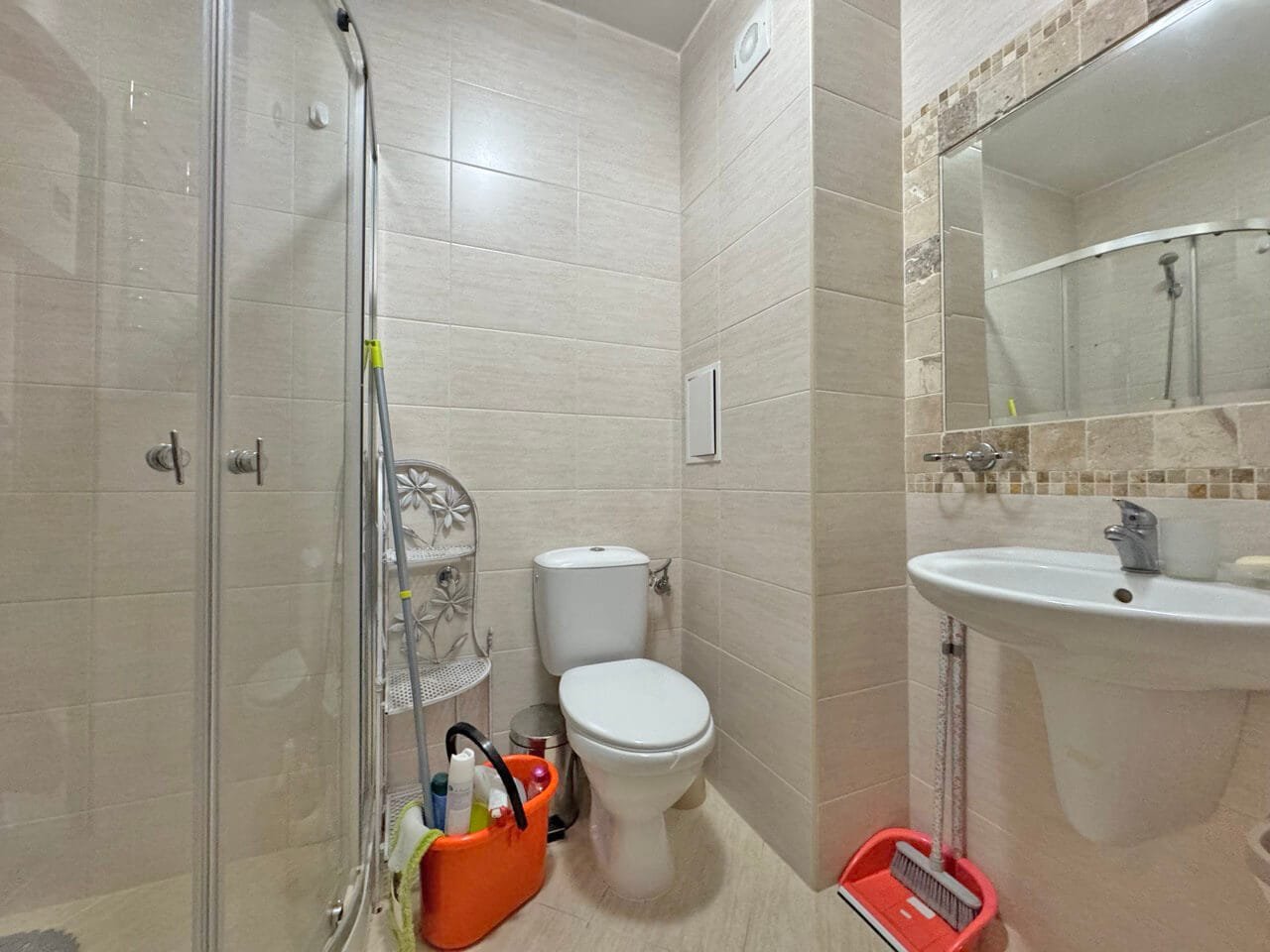 Современная ванная комната с душевой кабиной, унитазом, раковиной и уборочными принадлежностями в углу.