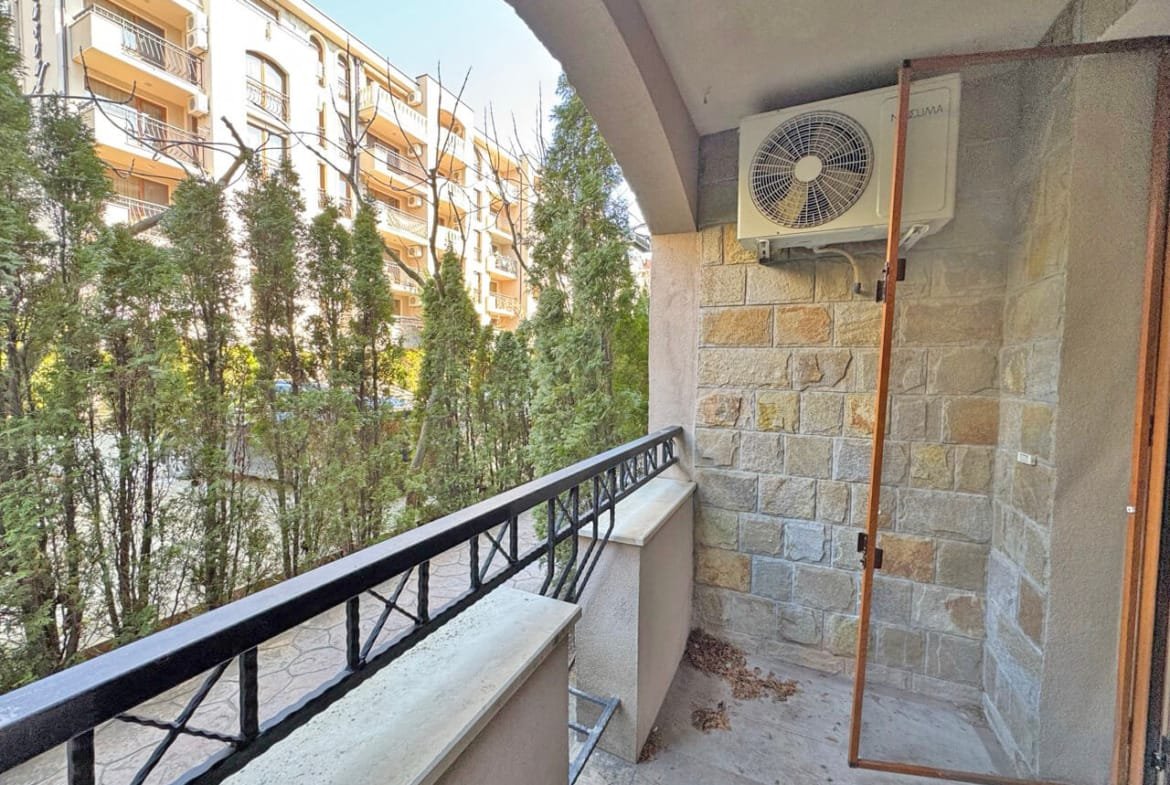 Балкон с кондиционером, окружённый зелёными кустами и видом на жилые здания. Яркое освещение и спокойная атмосфера.