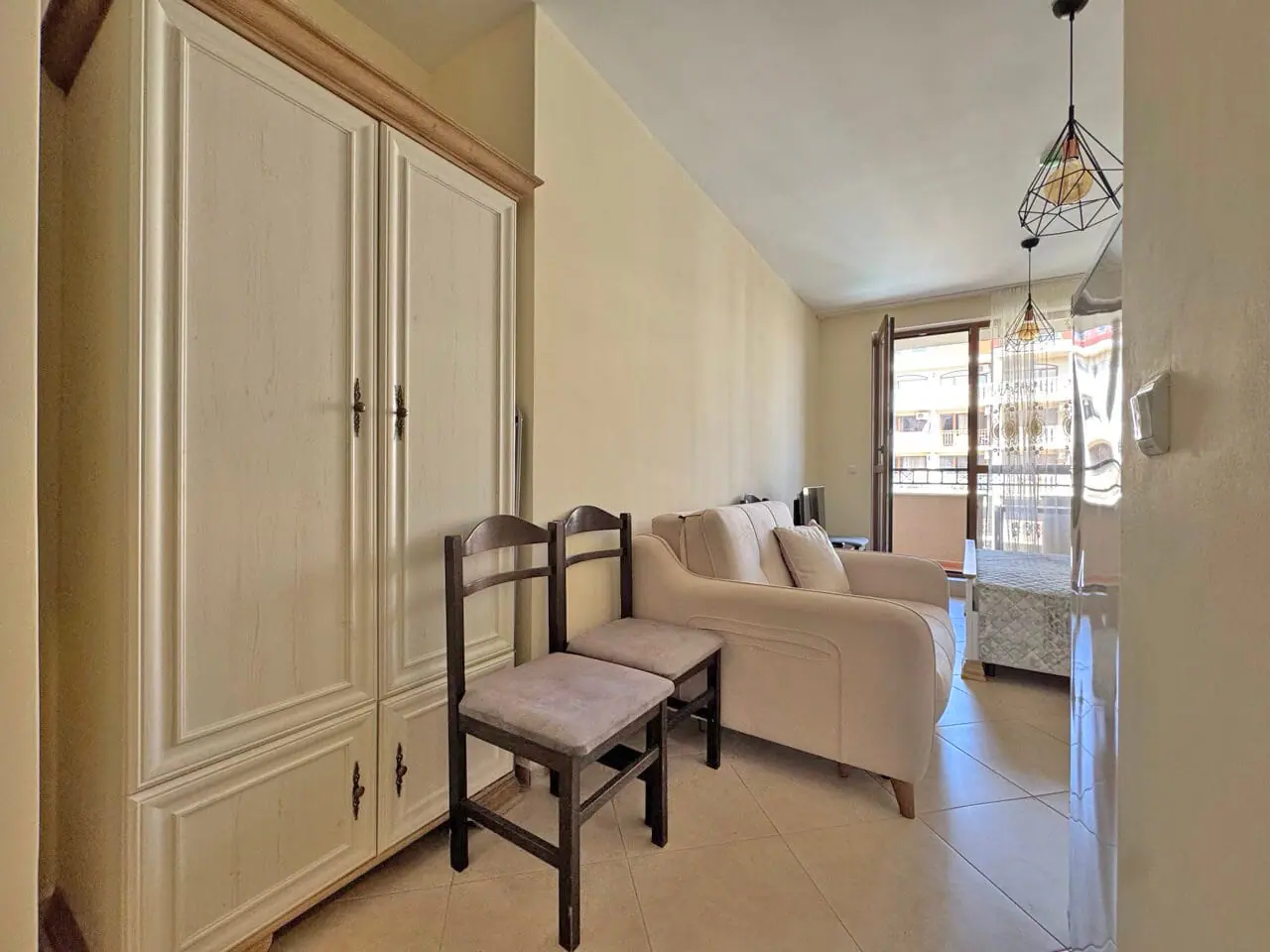 Гостиная с диваном и креслом, шкафом, большим окном и выходом на балкон, удобная для проживания в квартире.