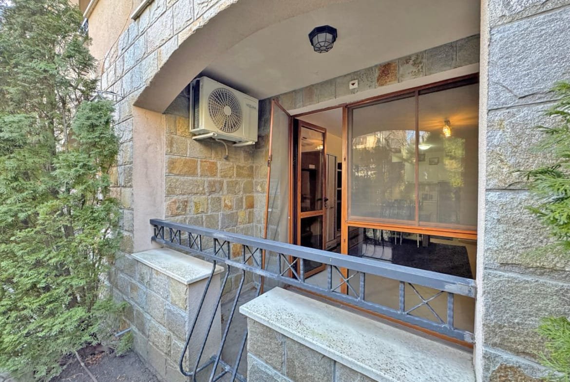 Открытая терраса с кондиционером и дверями к помещению, окруженная зелеными растениями и каменной стеной.