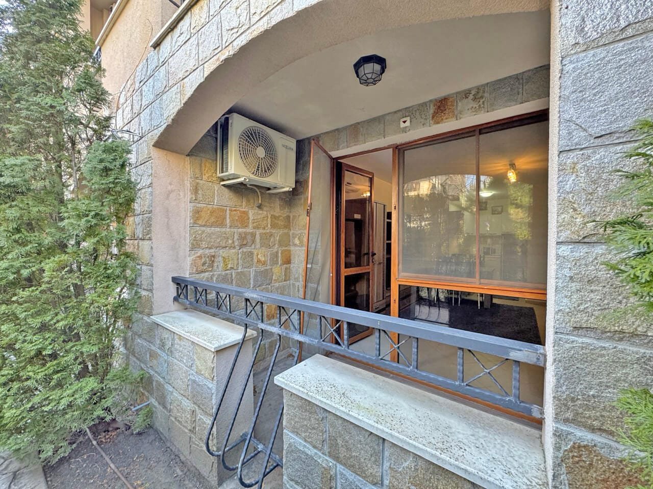 Открытая терраса с кондиционером и дверями к помещению, окруженная зелеными растениями и каменной стеной.