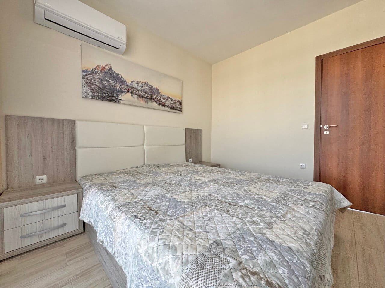 Современная спальня с деревянной мебелью, двуспальной кроватью и настенным искусством, светлые стены, холодные тона.