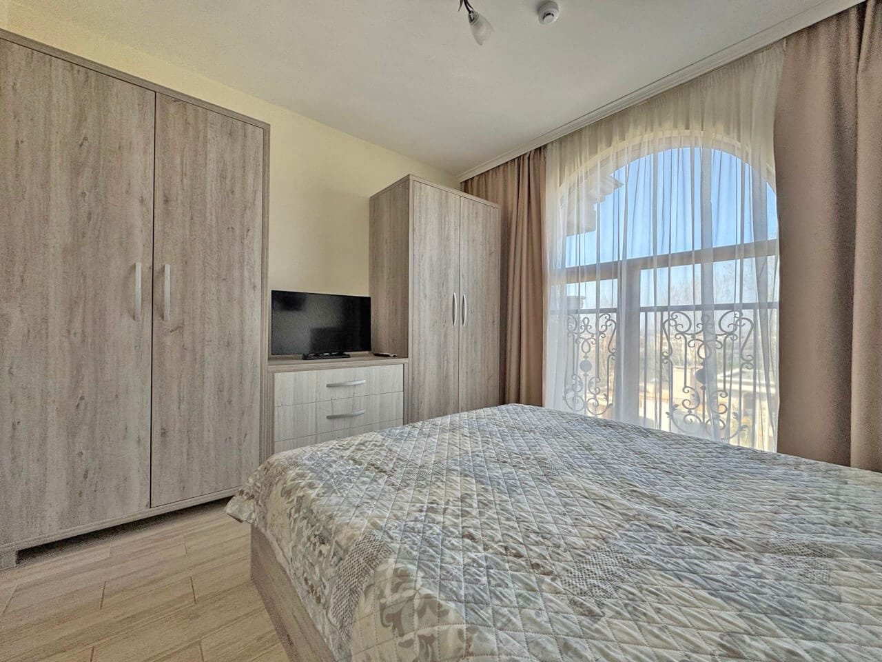 Уютная спальня с двойной кроватью, телевизором, шкафами и большим окном с занавесками.