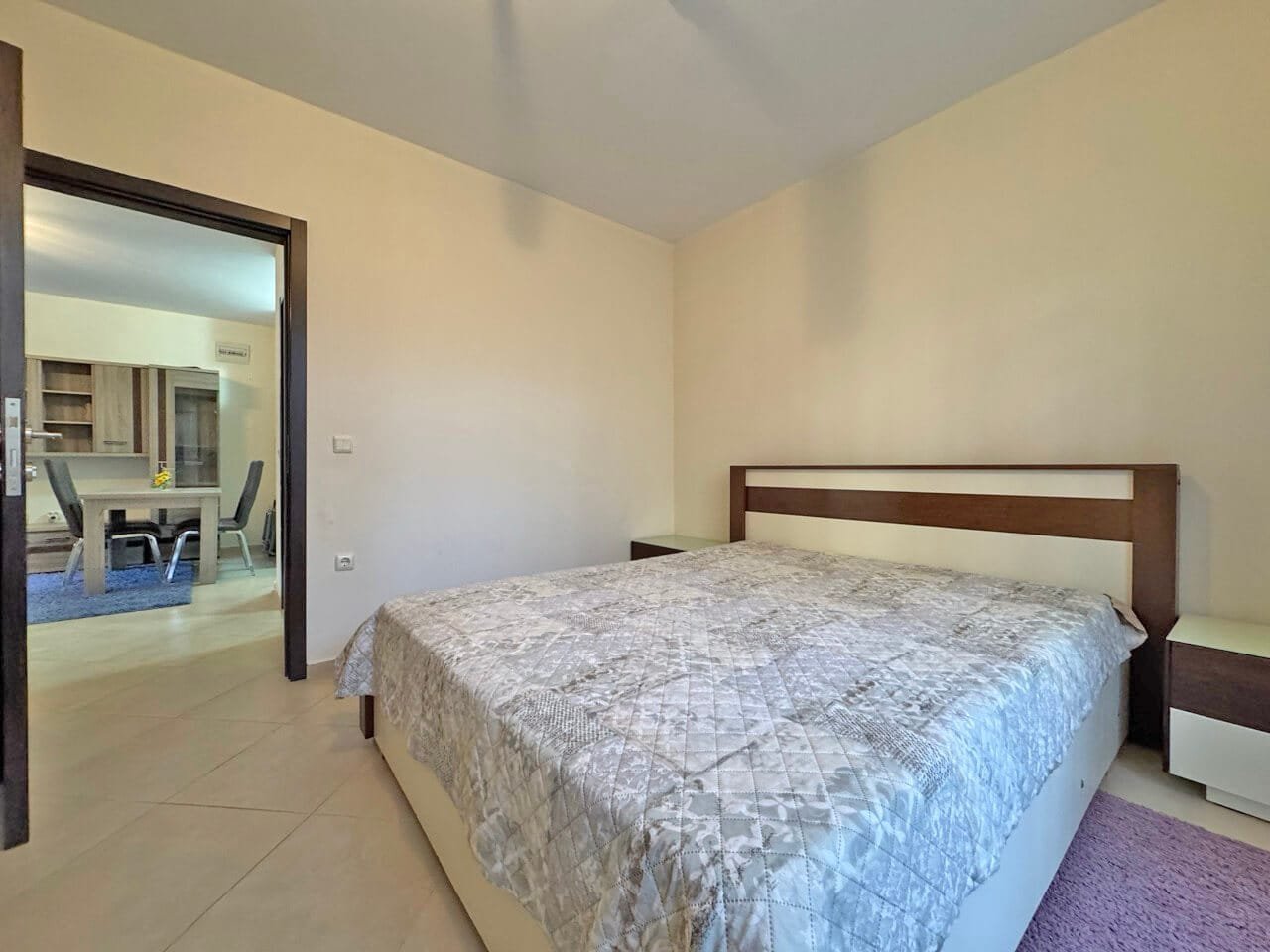 Спальня с двуспальной кроватью, светлыми стенами и уютным интерьером, вид на соседнюю комнату с обеденной зоной.