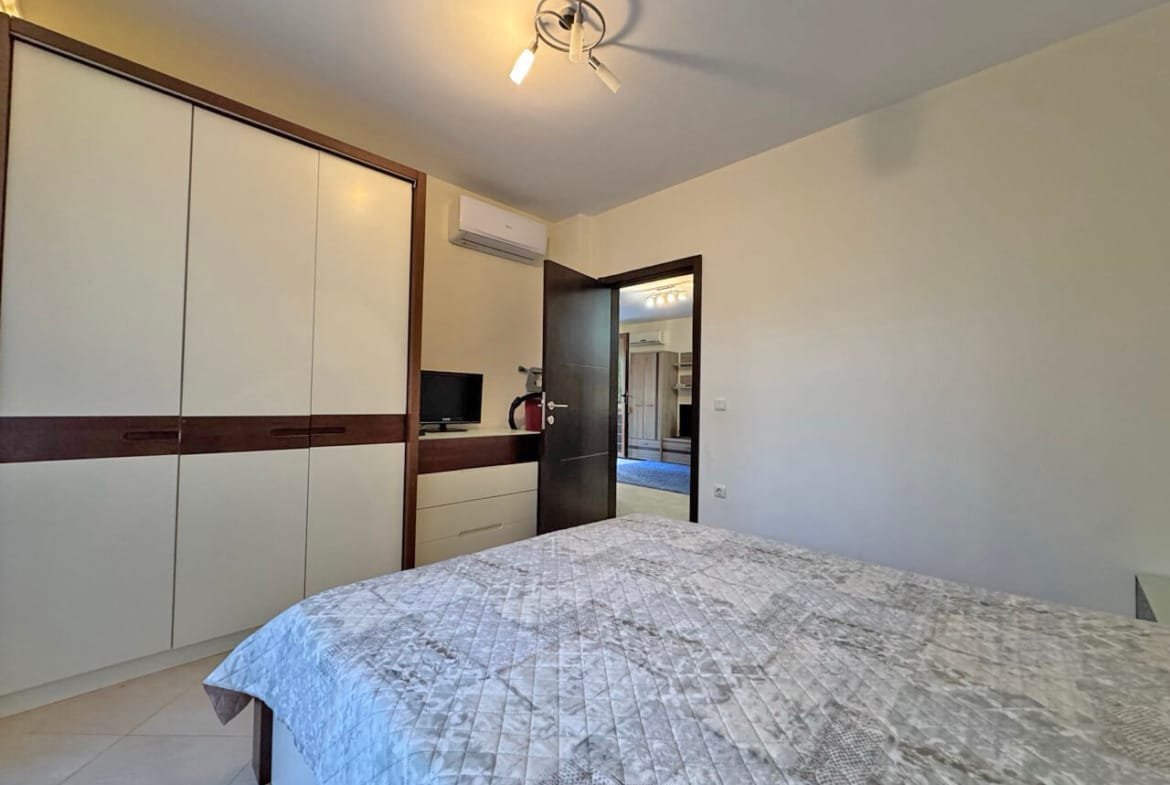 Современная спальня с двуспальной кроватью, шкафом, рабочим столом и мягким освещением. Стены светлые, пол плиточный.