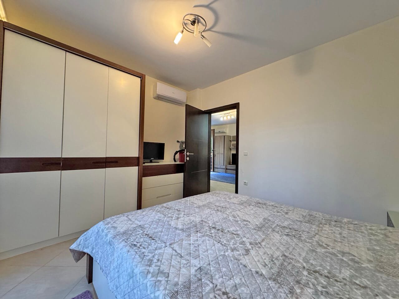 Современная спальня с двуспальной кроватью, шкафом, рабочим столом и мягким освещением. Стены светлые, пол плиточный.