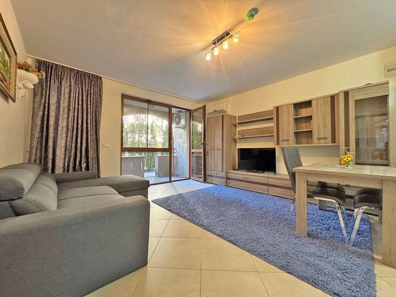 Современный интерьер квартиры с серым диваном, деревянными шкафами, телевизором и столом на фоне окон с зеленью.