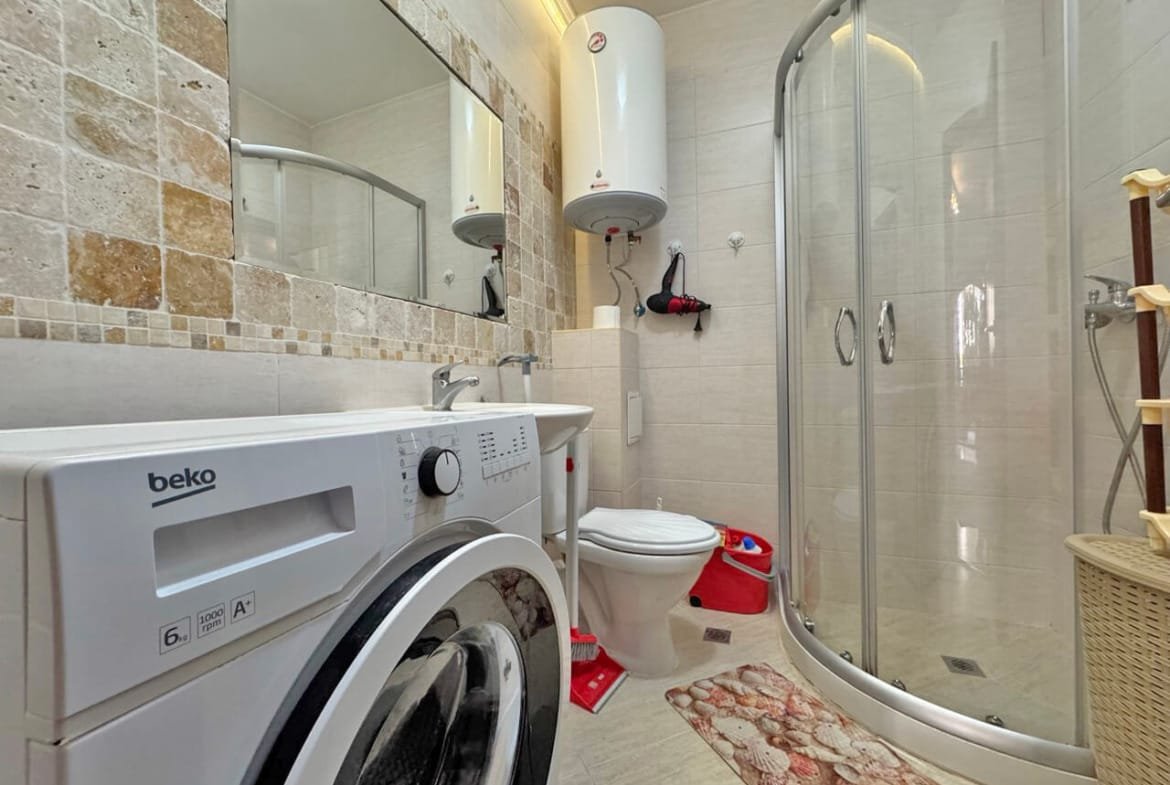 Современная ванная комната с душевой кабиной, стиральной машиной, унитазом и плиткой в бежевых тонах.