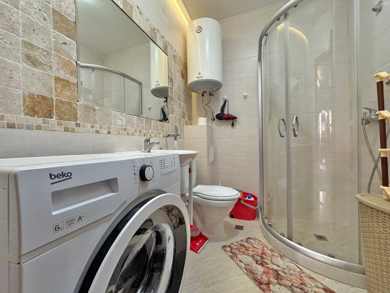 Современная ванная комната с душевой кабиной, стиральной машиной, унитазом и плиткой в бежевых тонах.