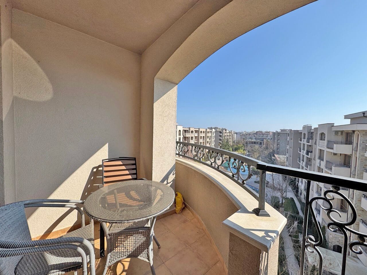 Балкон с круглым столом и стульями, overlooking жилые здания и ясное голубое небо.
