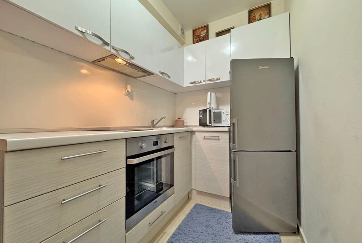 Современная кухня с белыми шкафами, серым холодильником, плитой, микроволновкой и ковриком на полу.