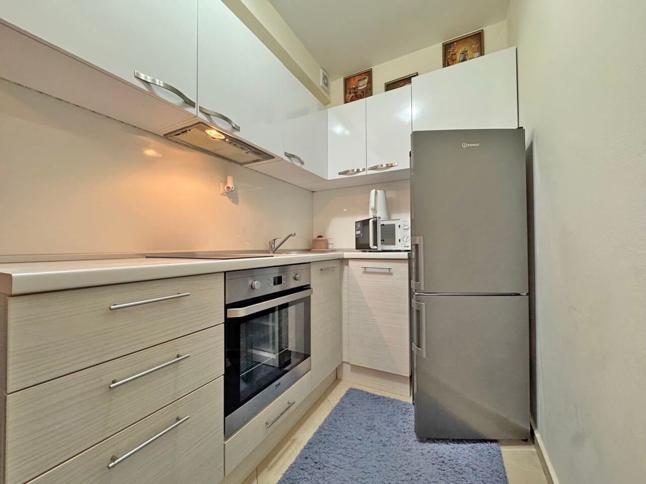 Современная кухня с белыми шкафами, серым холодильником, плитой, микроволновкой и ковриком на полу.