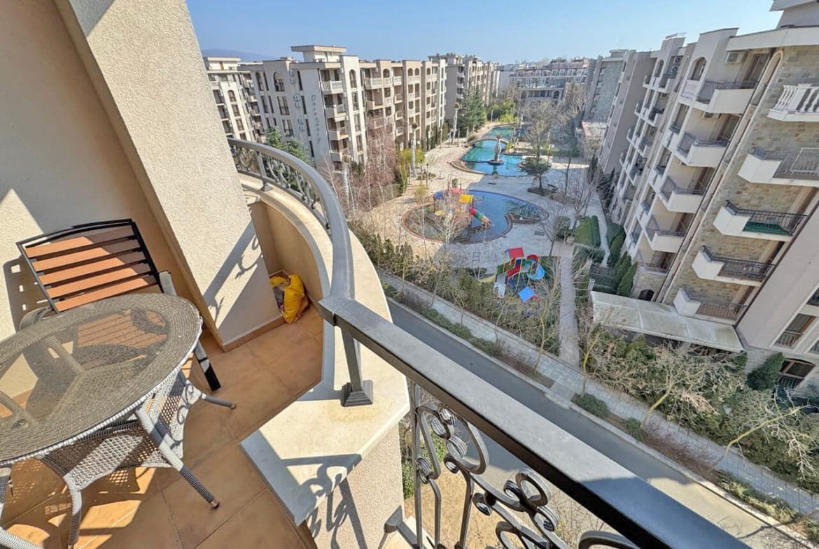 Балкон с креслом и столом, видом на ухоженный двор с бассейном и детскими площадками, окруженный жилыми домами.