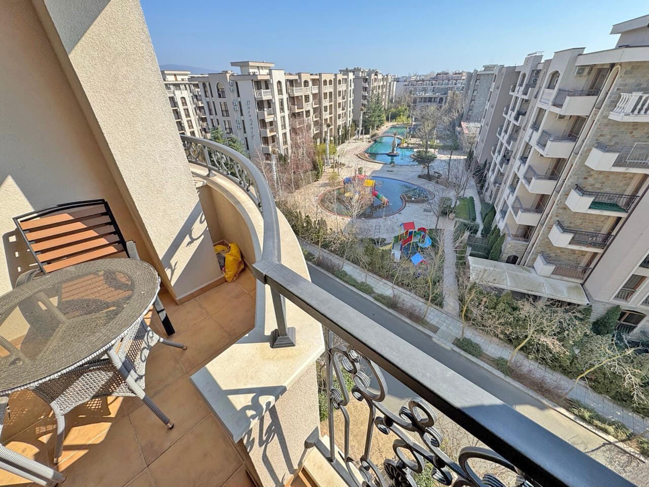 Балкон с креслом и столом, видом на ухоженный двор с бассейном и детскими площадками, окруженный жилыми домами.