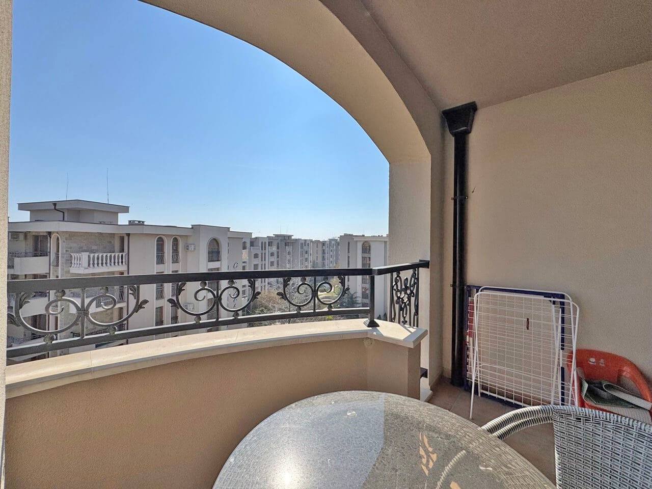 Балкон с металлическим ограждением, столом и стульями, с видом на современные здания и ясное небо.